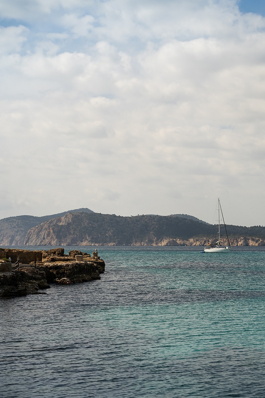 Rocky coastline meets turquoise Mediterranean waters with a white sailboat anchored before distant, arid hills.