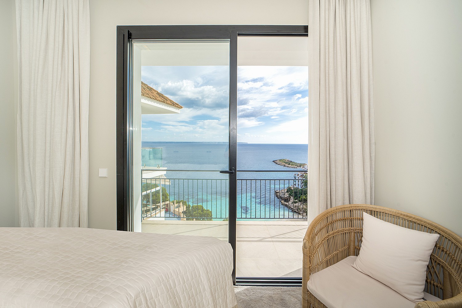 Interior view of a bedroom featuring sliding glass doors opening to a sea-view balcony with a woven chair in the foreground.