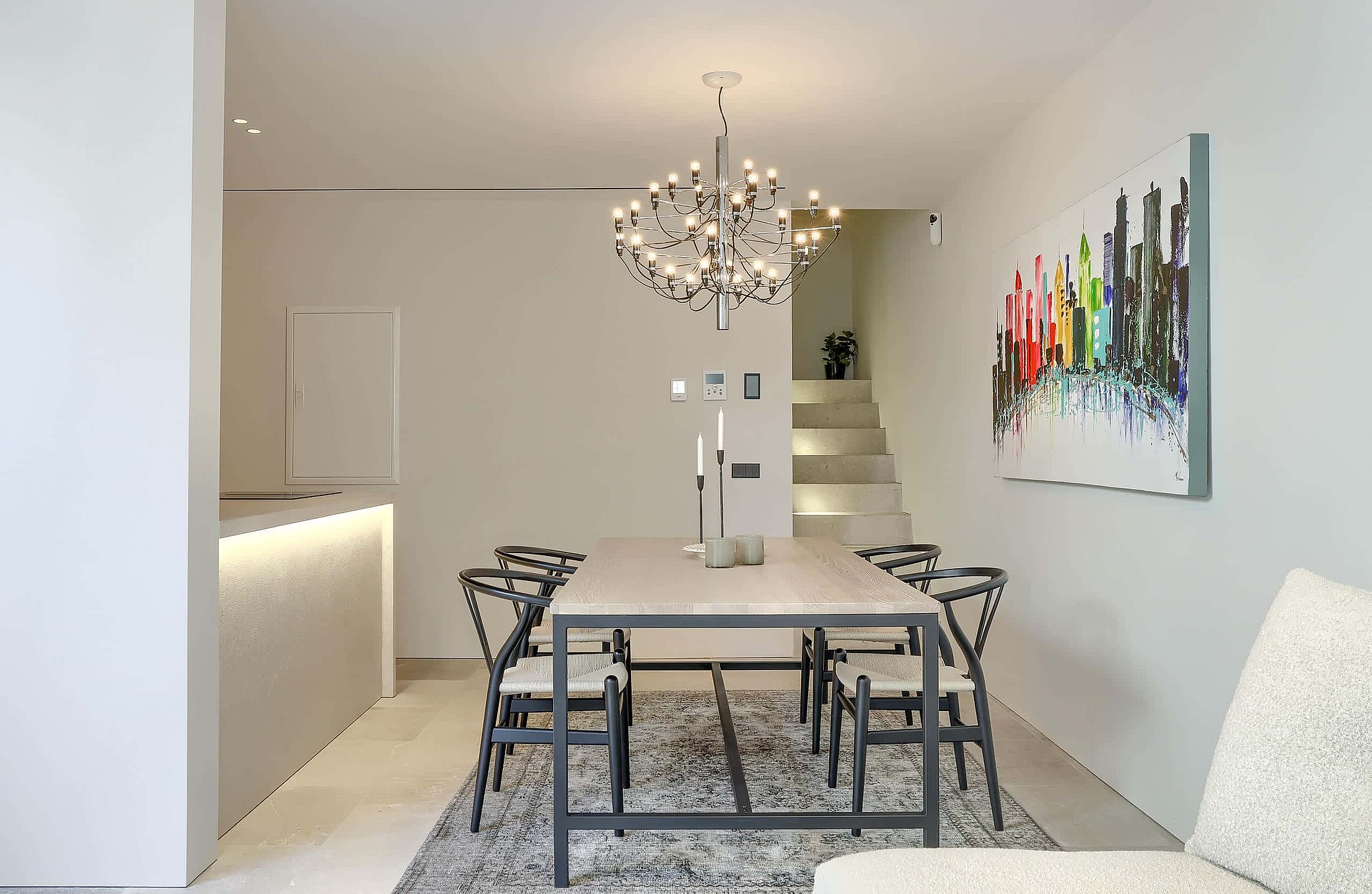 Interior view of a minimalist dining room with a stone table, black wooden chairs, a modern branching chandelier, and an abstract multi-colored painting on a white wall.