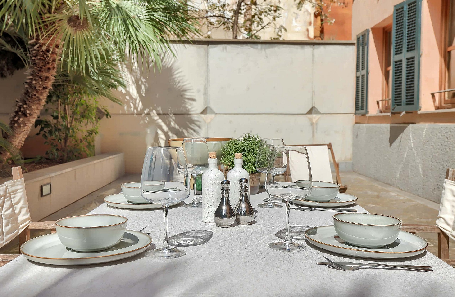A dining table set for four on a stone terrace featuring cream-colored linens, ceramic bowls, and palm tree shadows.