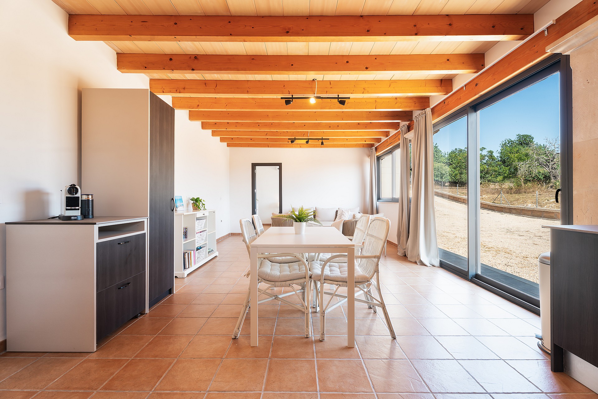 Comedor con mesa y sillas blancas, suelos de baldosas y puertas correderas hacia el exterior.