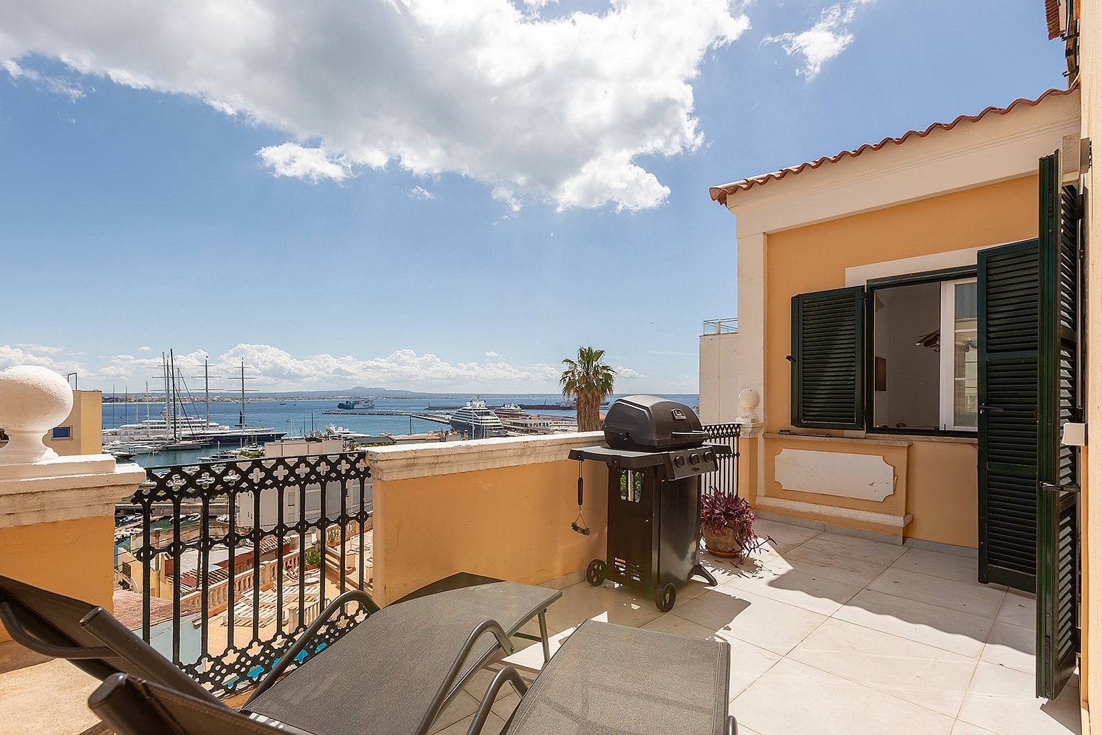 A sunlit Mediterranean terrace featuring wrought-iron railings, lounge chairs, a barbecue grill, and open views of a marina.
