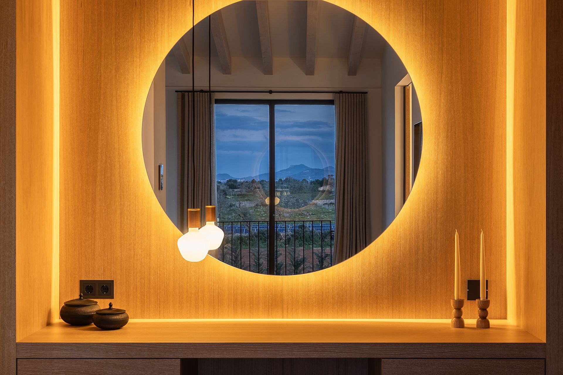 A backlit circular mirror over a wooden vanity table, reflecting a window with dusk mountain views in a Santanyí finca.