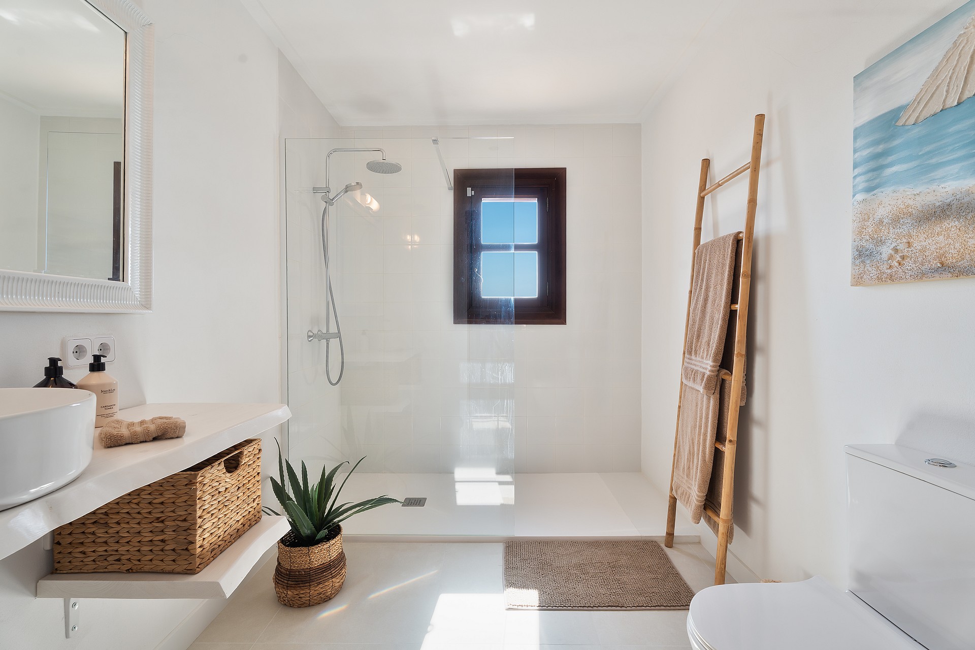 Baño blanco con ducha a ras de suelo, toallero de madera y detalles en fibras naturales.