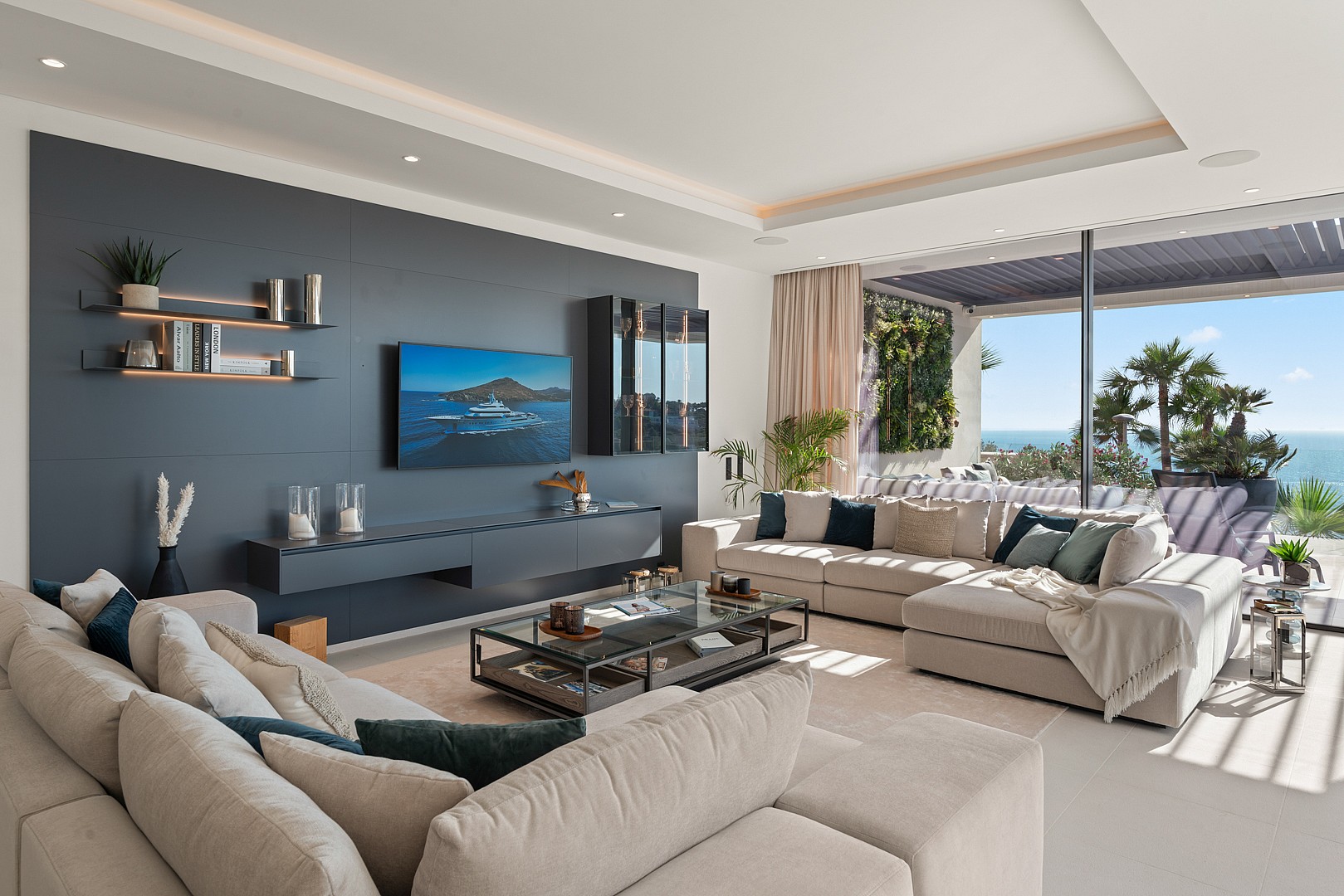 Modern living room with a charcoal grey feature wall, a wall-mounted flat-screen television, a beige sectional sofa, and floor-to-ceiling glass sliding doors opening to a terrace with sea views.
