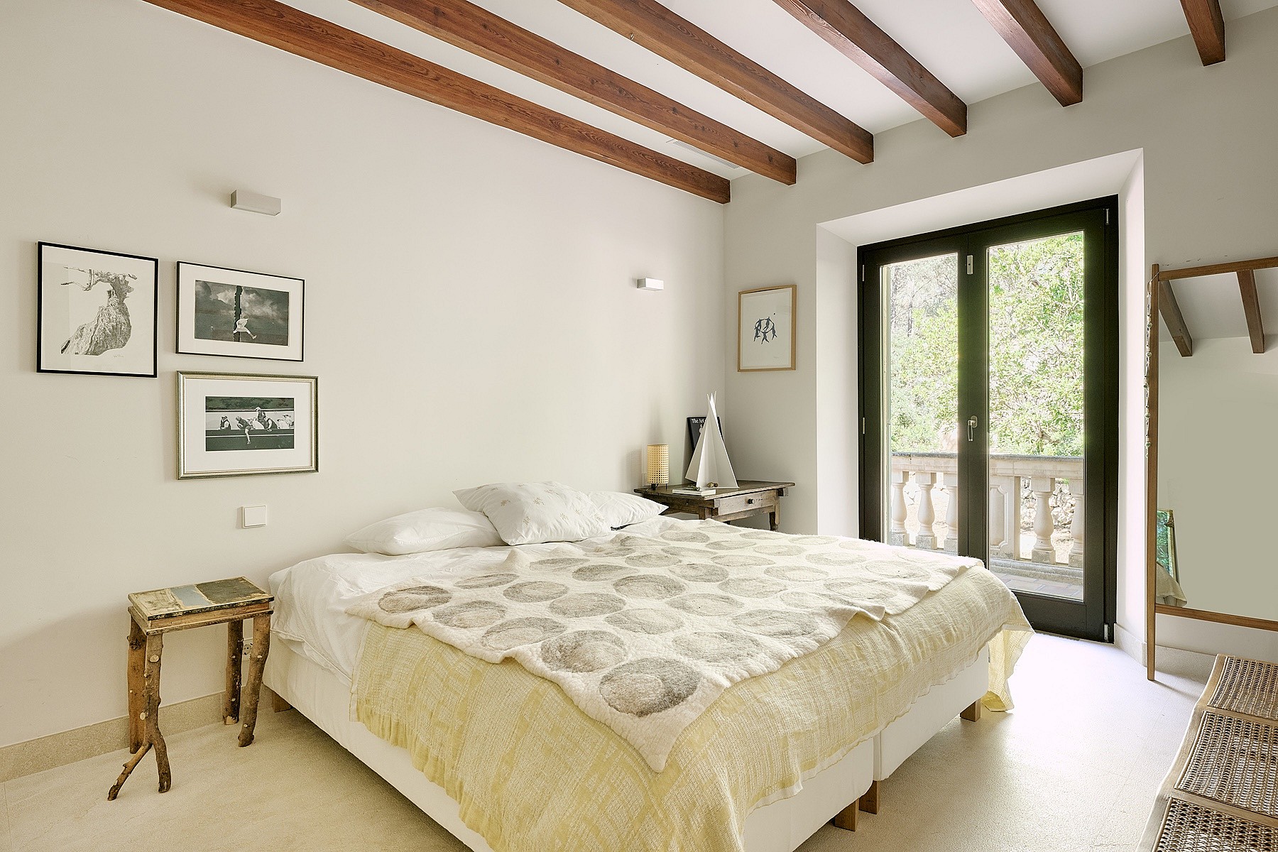 A bright bedroom with a queen bed, exposed dark wood ceiling beams, cream walls, stone flooring, and glass doors to a balcony.