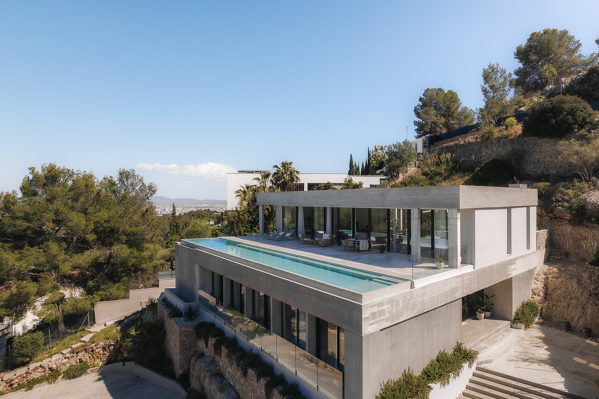 Elevated view of a contemporary concrete villa featuring a rooftop infinity pool and floor-to-ceiling glass walls.