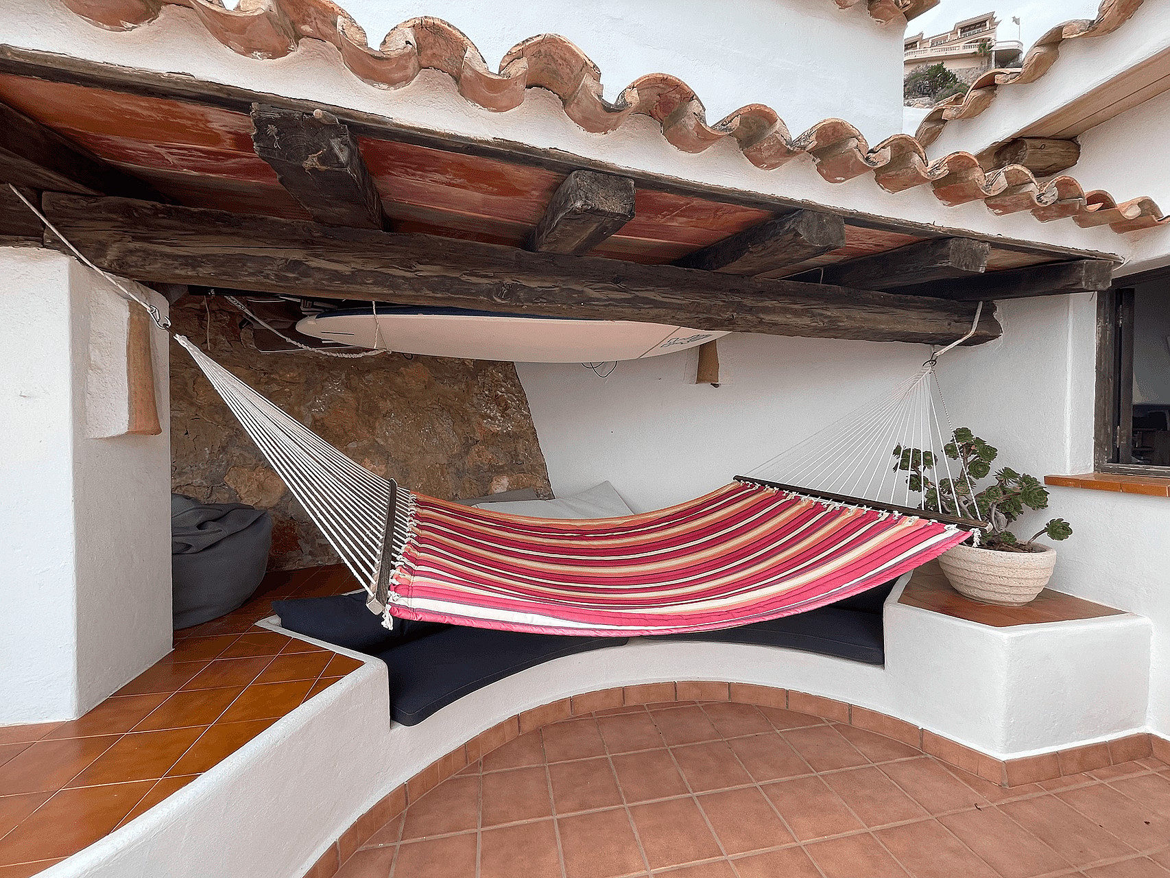 A covered terrace nook featuring a striped fabric hammock strung between white walls under a rustic wooden beam roof.