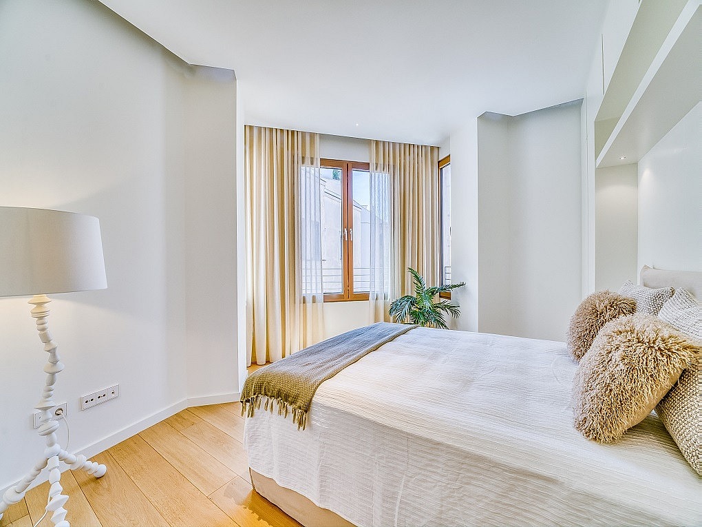 A bright bedroom with hardwood floors, a white bed with textured pillows, and beige floor-to-ceiling curtains.