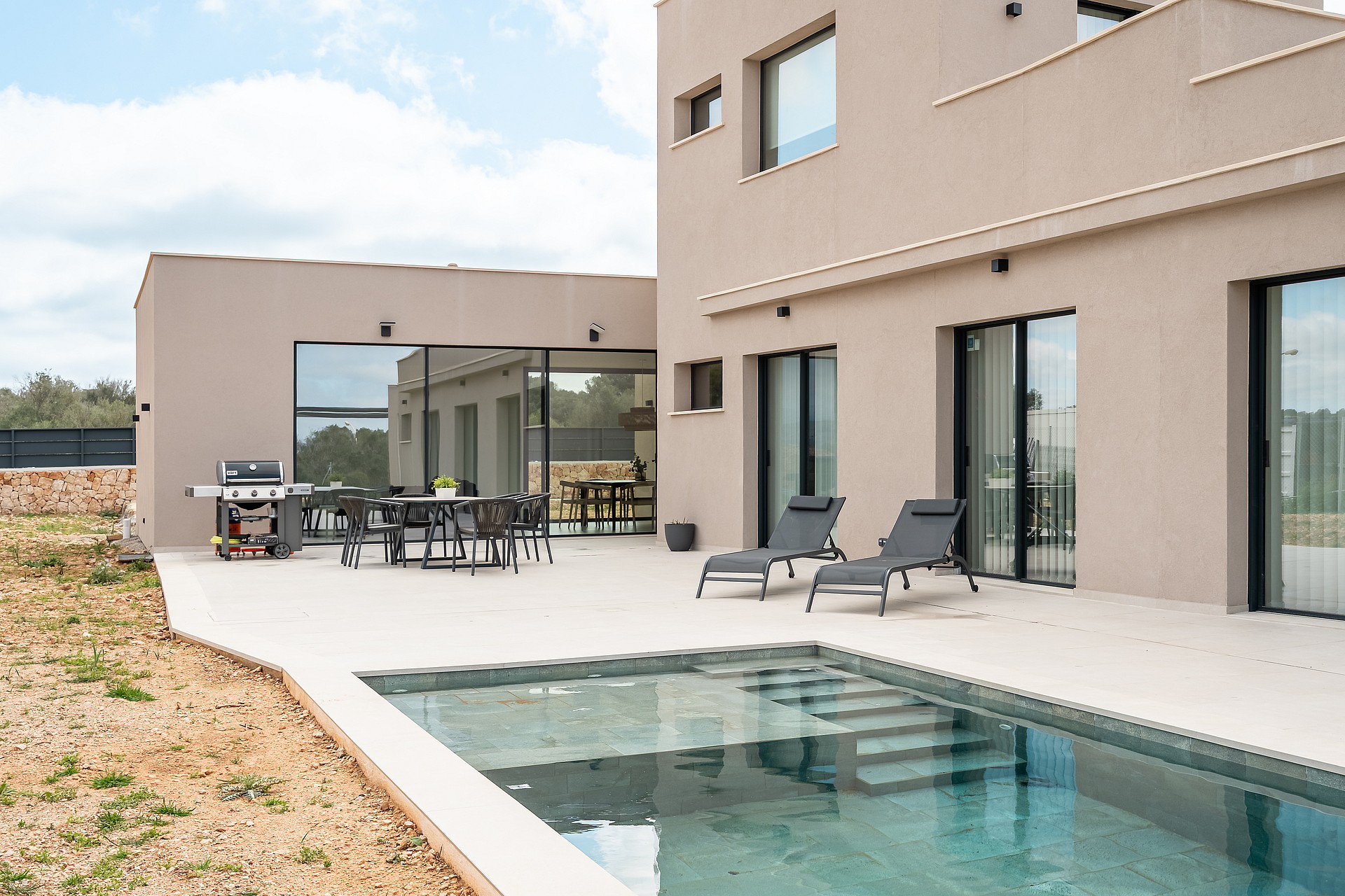 Rear facade of a modern beige villa showing a stone terrace, swimming pool with steps, and outdoor dining area.