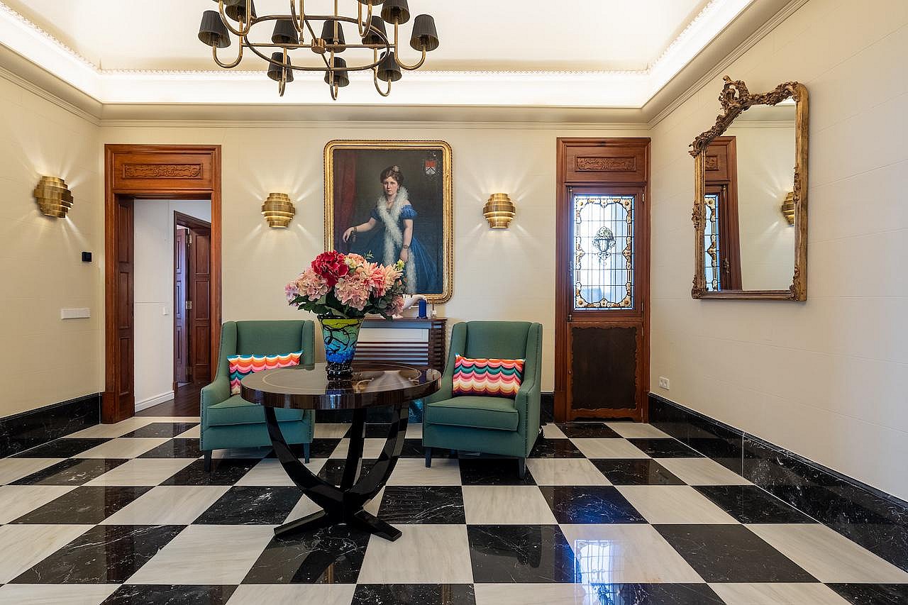 A formal entrance hall featuring black and white checkered marble floors, two teal armchairs, and a round central pedestal table.