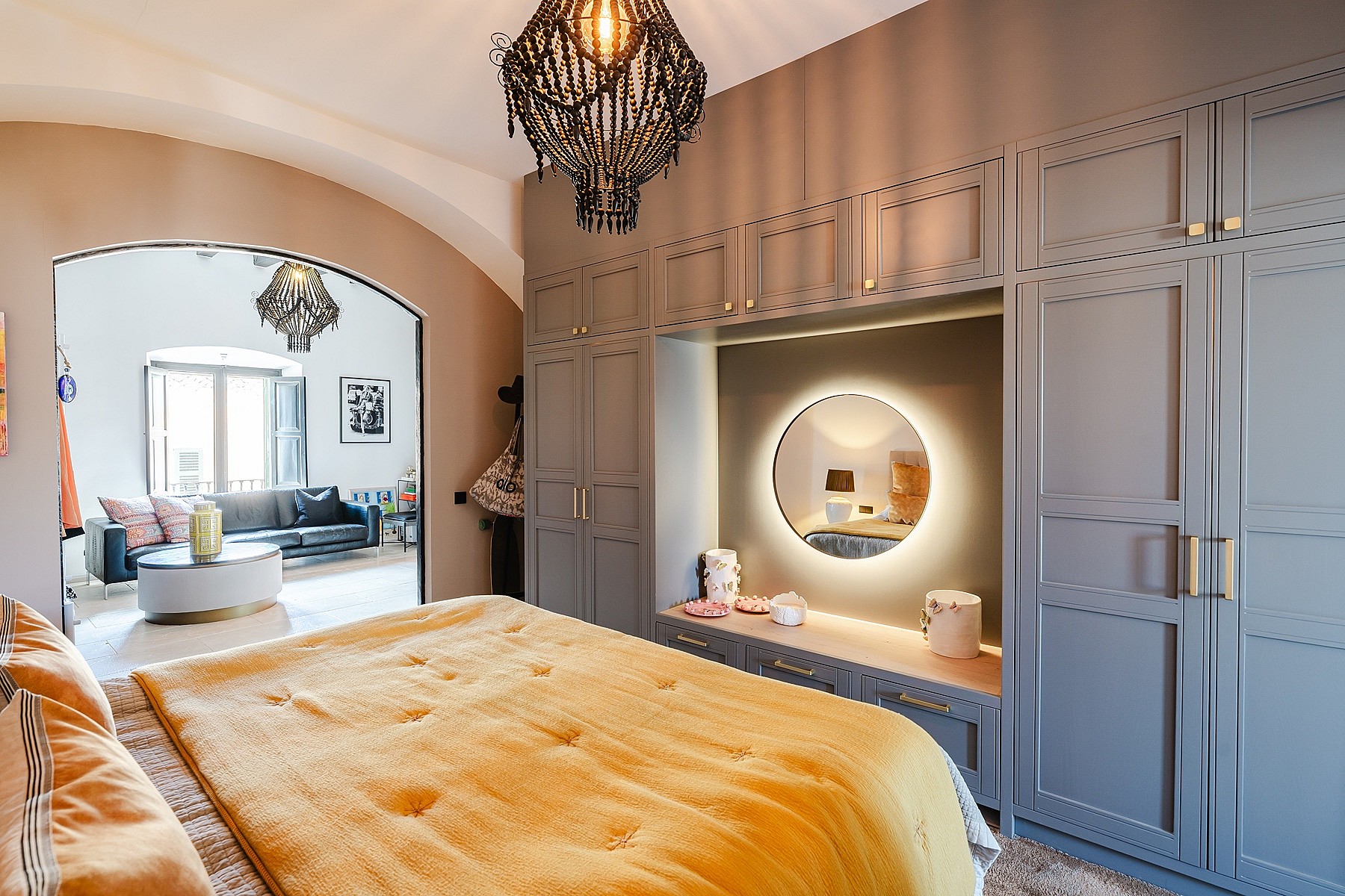 A master bedroom featuring a large bed with a golden throw, grey paneled wardrobes, a circular backlit mirror, and an arched doorway leading to a sitting area.