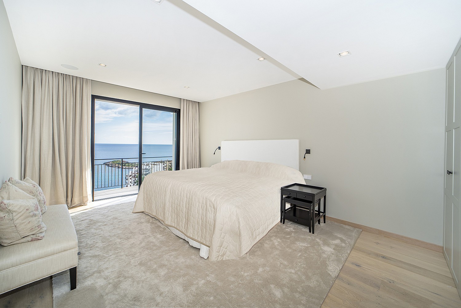 A minimalist bedroom featuring a large bed, neutral carpeting, light grey walls, and floor-to-ceiling glass sliding doors opening to a sea-view balcony.
