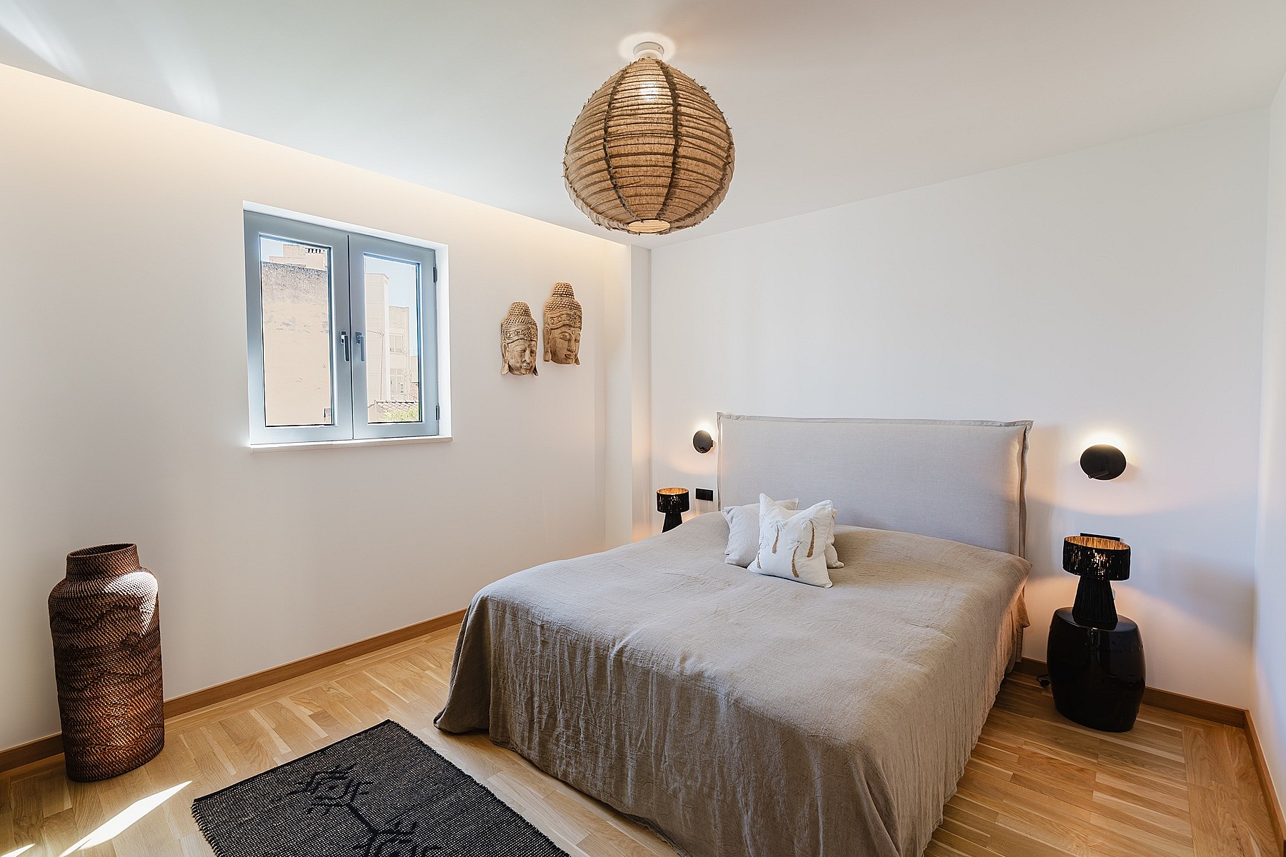 A minimalist bedroom featuring a grey upholstered headboard, light oak wood flooring, and a large woven pendant light.