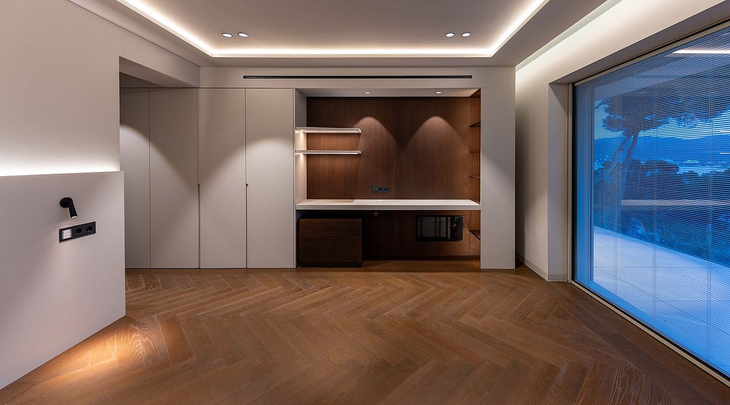 A modern home office space featuring dark wood herringbone flooring, integrated cabinetry with white countertops, and expansive glass sliding doors.