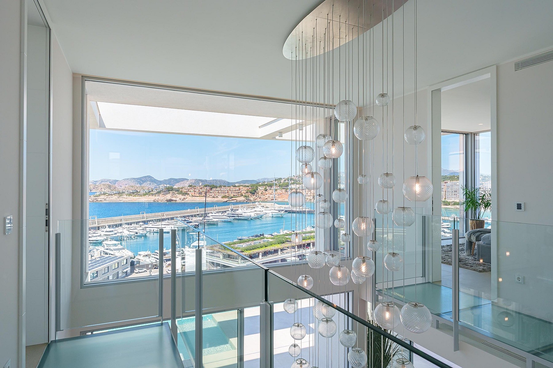 Top of a glass staircase featuring a cluster pendant light, with a wide window framing a view of Port Adriano harbor.
