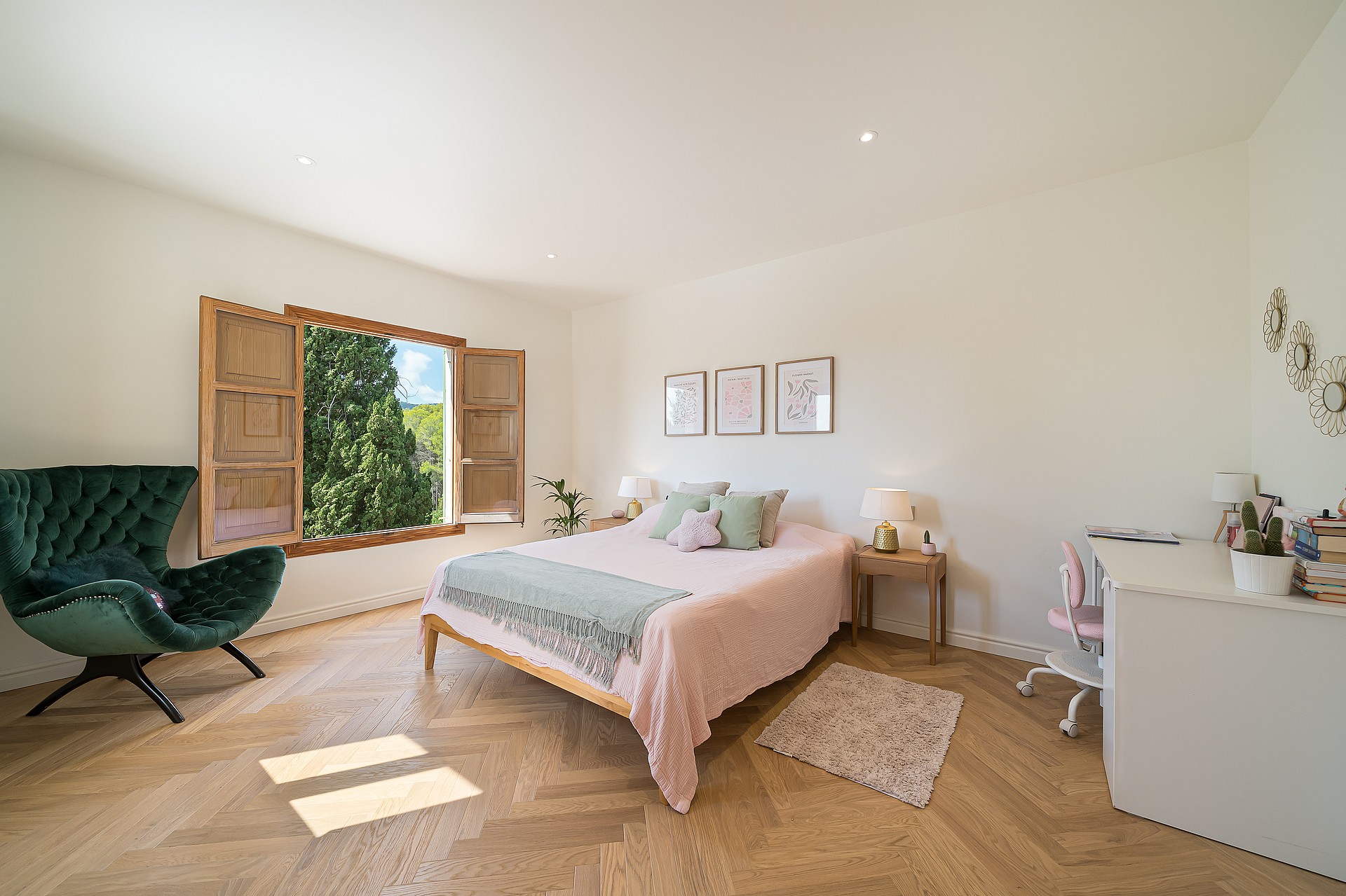 A bright bedroom featuring light oak herringbone parquet, a green velvet armchair, and wooden shutters opening to trees.