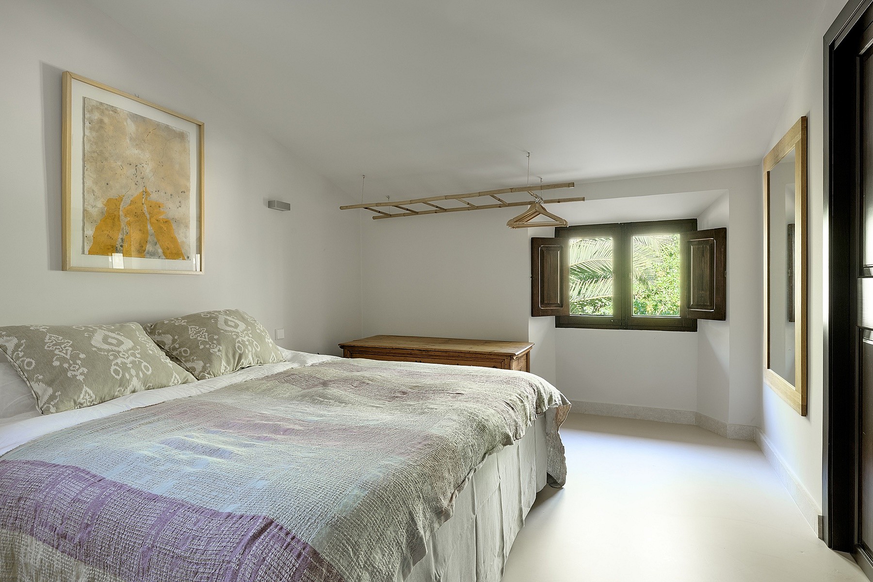 A bright bedroom in a finca with white walls, a bed featuring a patterned spread, a wooden chest, and an open casement window.