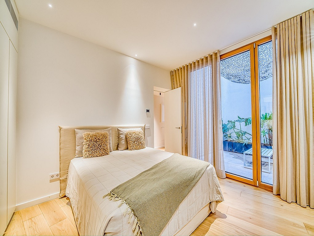 A modern bedroom with light wood floors, a neutral bed, and floor-to-ceiling glass doors opening to a private patio.