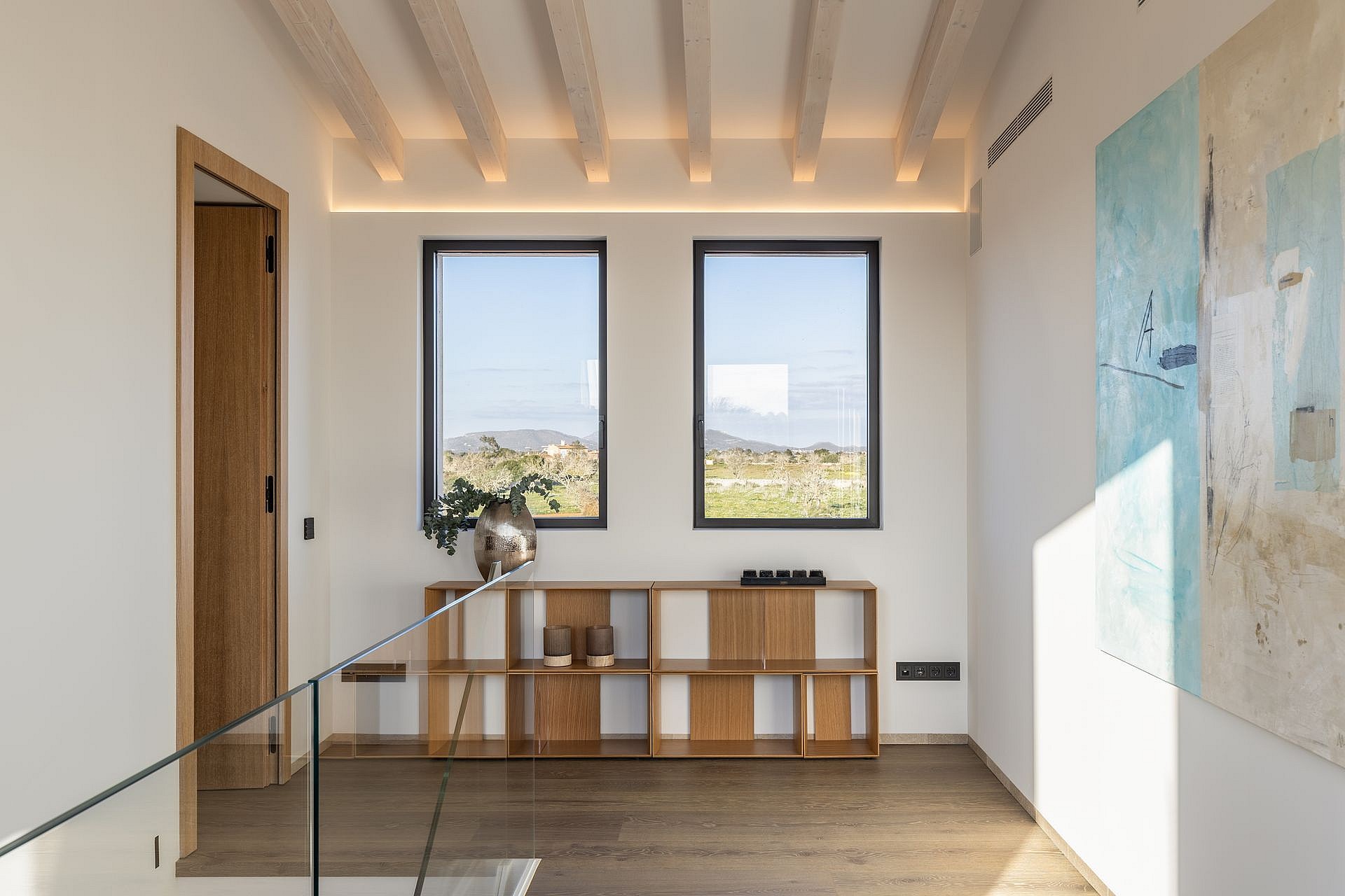 A bright hallway featuring wooden flooring, a low timber bookshelf, white walls with an abstract painting, and two windows showing mountain scenery.