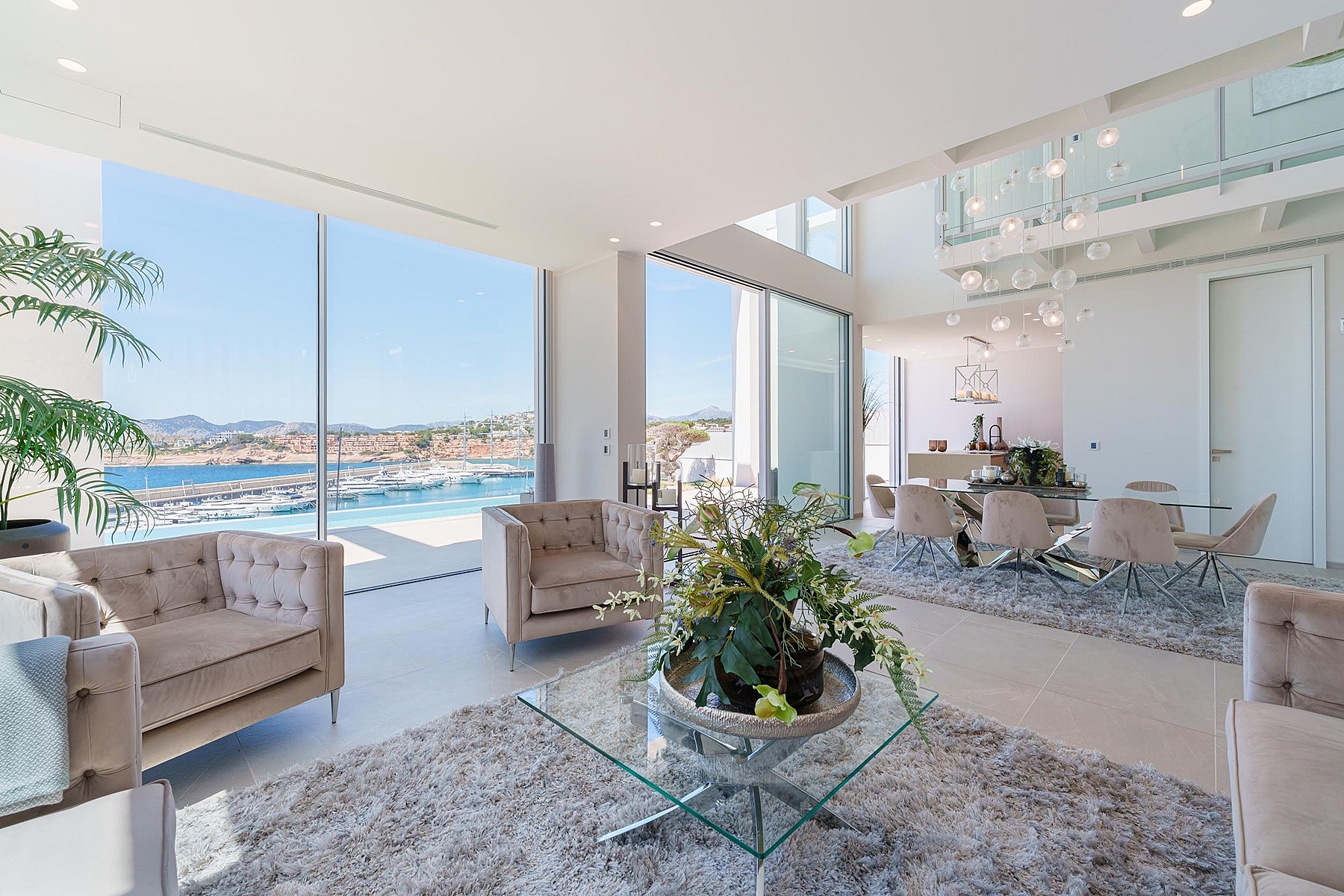 Modern open-plan living room with floor-to-ceiling windows overlooking Port Adriano marina, featuring tufted furniture.