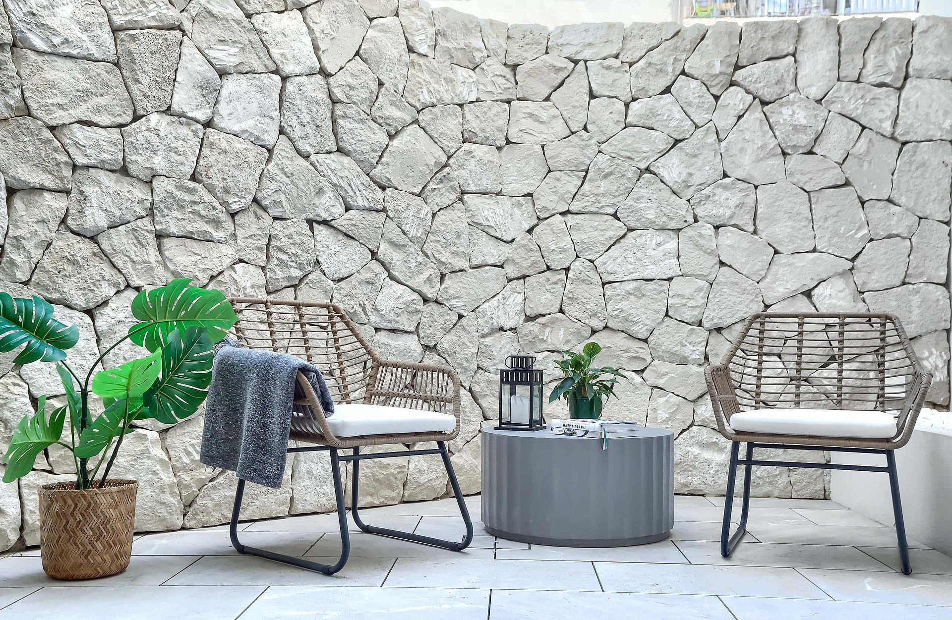 A small outdoor seating area featuring two wicker chairs with white cushions and a grey round side table against a tall, natural dry-stone wall.
