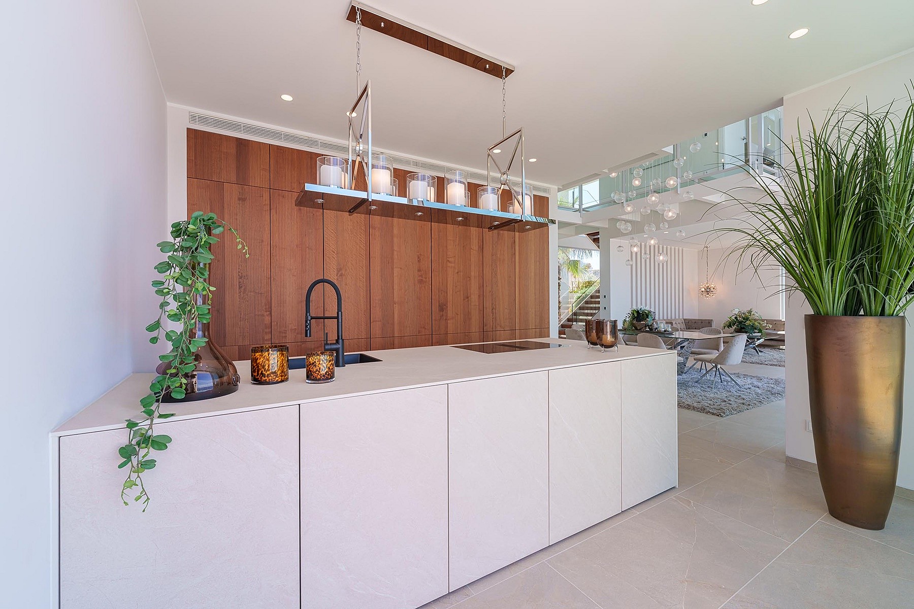 Modern kitchen island with matte white surfaces and dark wood wall-to-ceiling cabinetry, interior glass staircase visible in background.