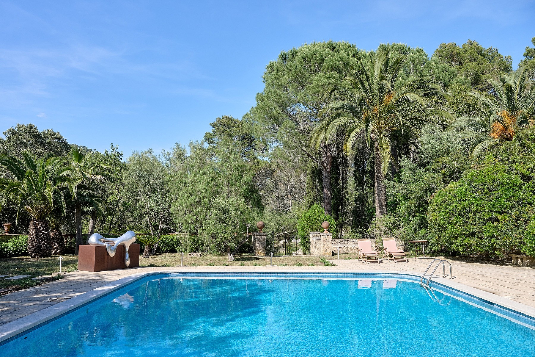 A rectangular swimming pool with blue water sits in a landscaped garden, surrounded by trees, palm trees, and gravel paths.