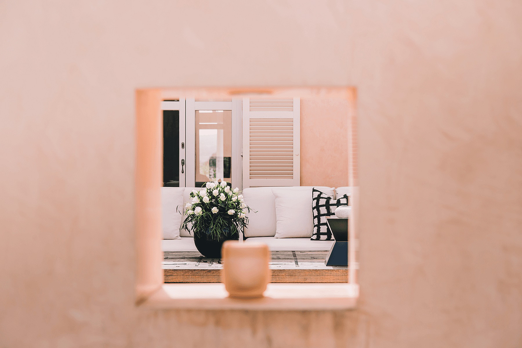 A square architectural opening in a pink stucco wall reveals a white sofa with patterned pillows and a potted plant on a terrace.