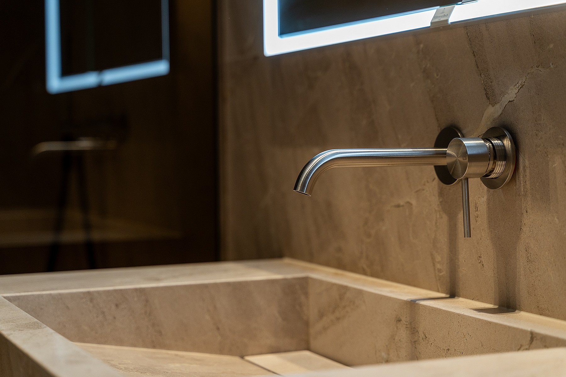 Close-up of a minimalist stone sink with a chrome wall-mounted faucet and backlit mirror in a modern bathroom.
