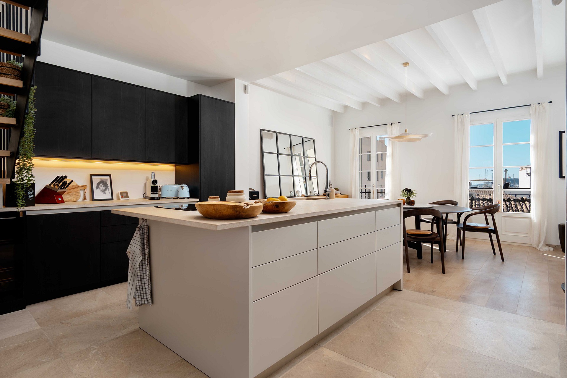 Open kitchen with black cabinetry, a pale stone-topped island, and a dining table and French doors visible beyond.