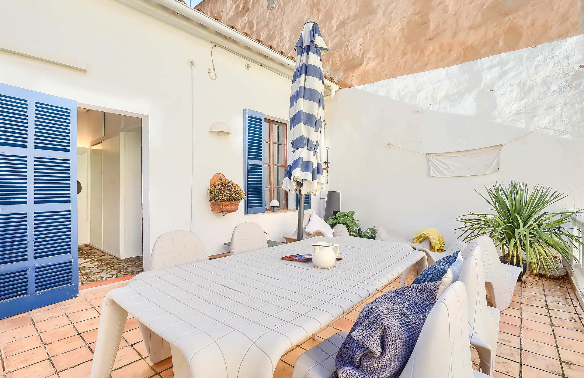 Outdoor terrace with a dining table and chairs, blue-striped sun umbrella, and blue shutters on a white building exterior.
