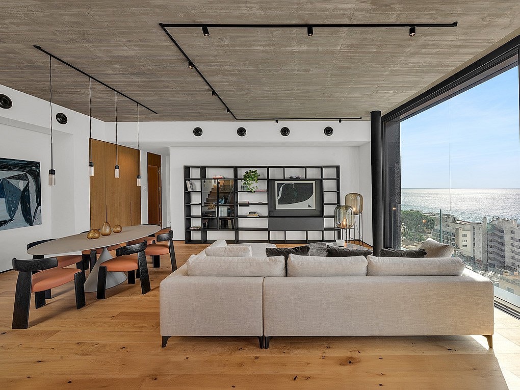 Wide angle interior view of an open-plan living room with light oak floors, raw concrete ceiling, and black track lighting.