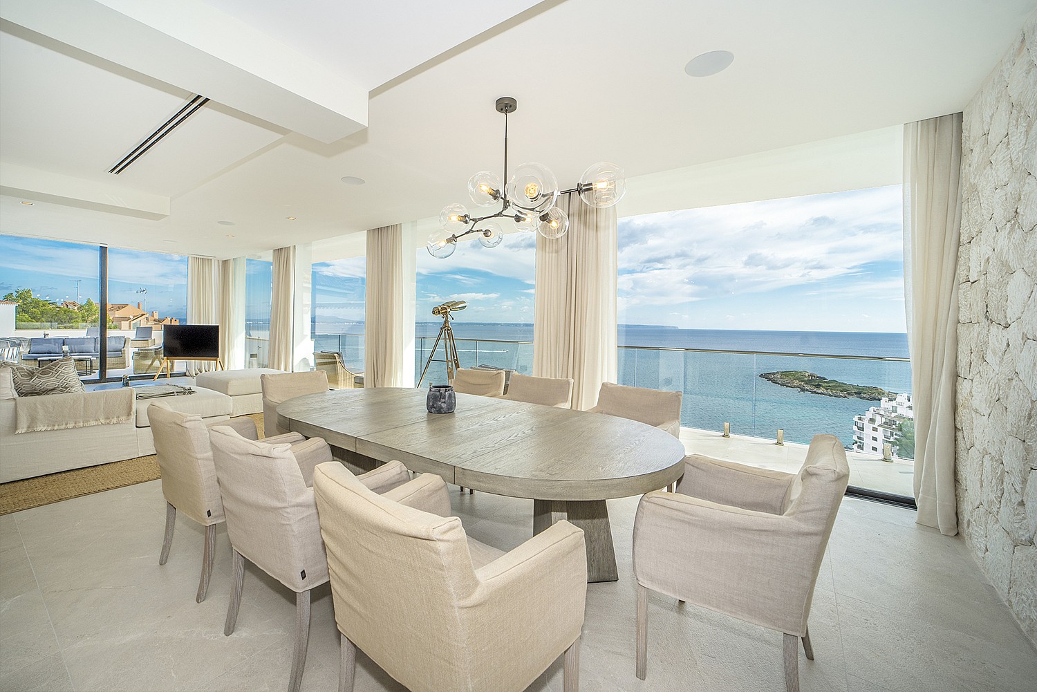 A dining area with an oval stone table and beige chairs, featuring a sculptural chandelier and a large glass-walled balcony.