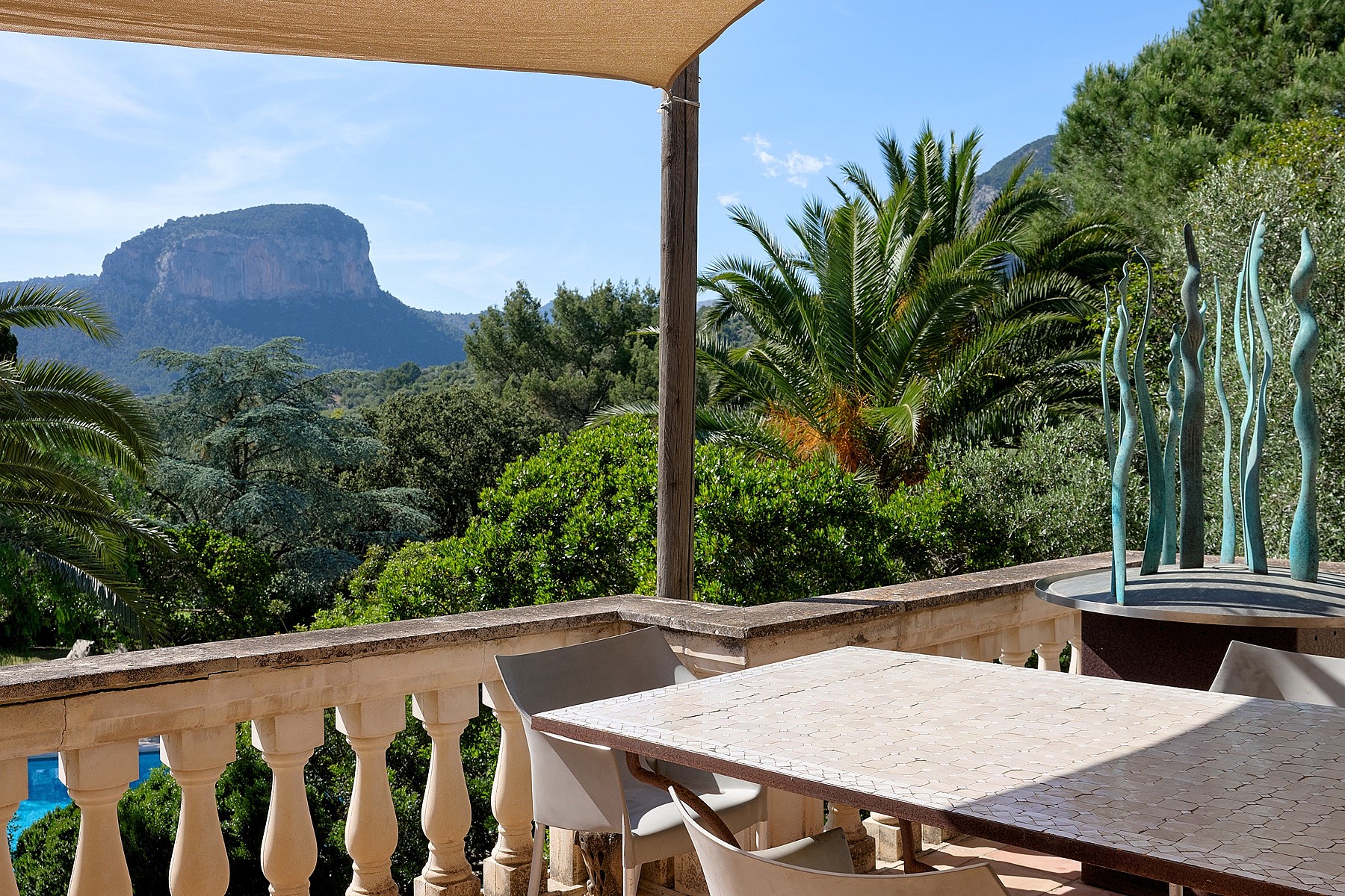 An outdoor terrace with a stone balustrade, a stone dining table, and a shaded seating area overlooking Alaro mountain peaks.