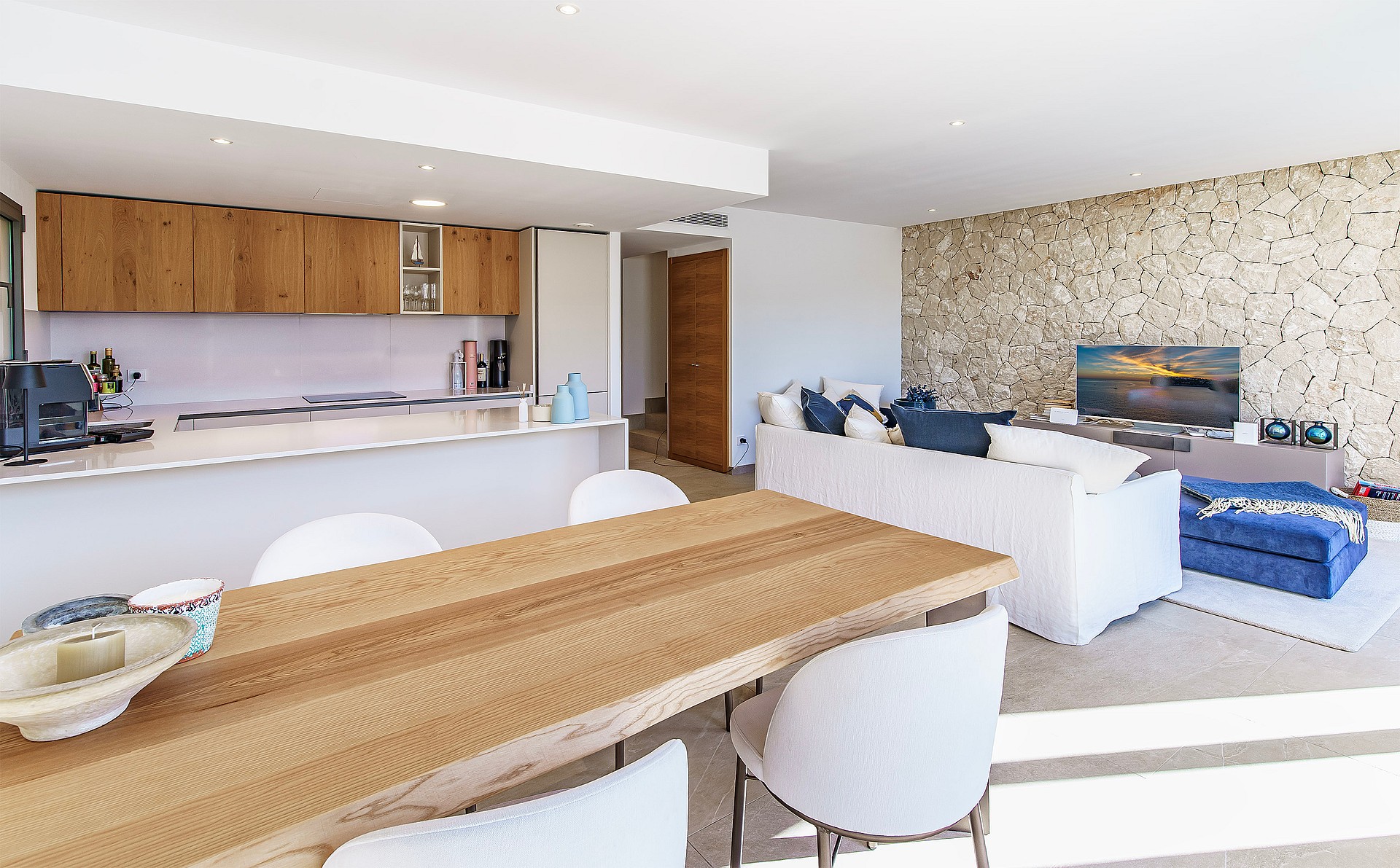 A bright open-plan space showing a kitchen with wooden cabinetry, a breakfast bar, and a living area featuring a stone wall.