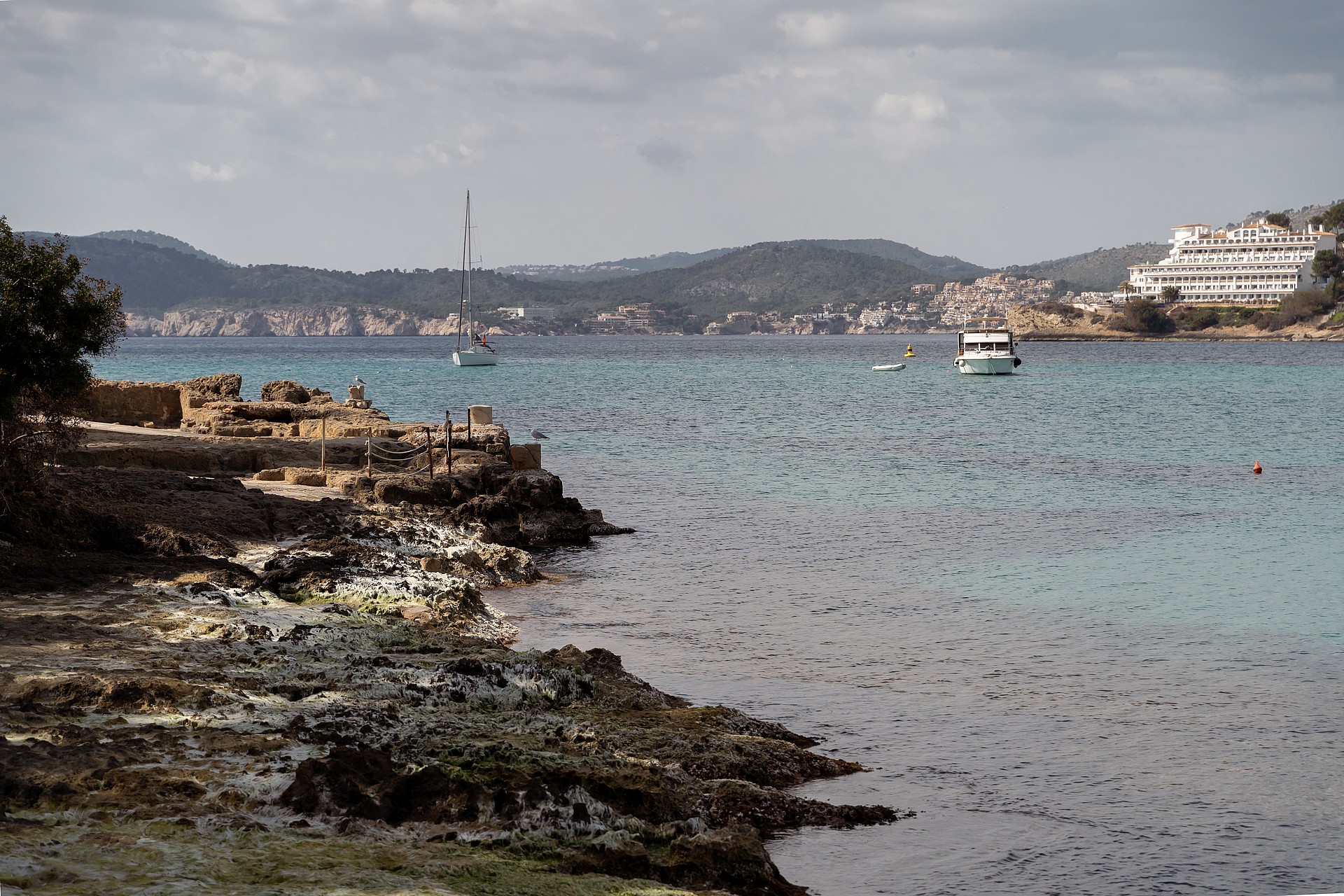 Rocky Mediterranean shoreline with turquoise water, a distant sailboat, and coastal hills under an overcast sky.