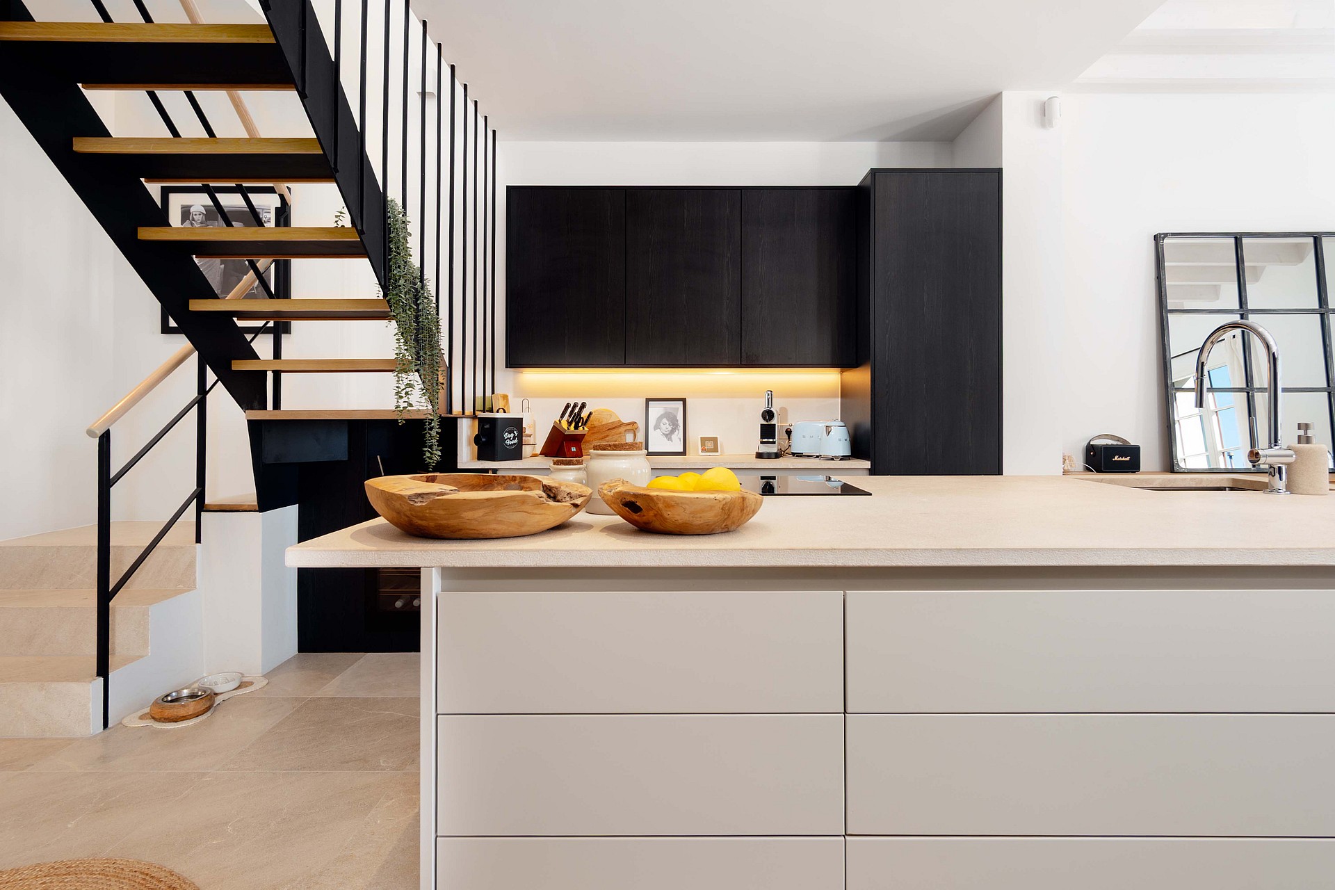 Modern kitchen with matte black cabinetry, stone-topped island, and an open-tread metal and wood staircase in the foreground.