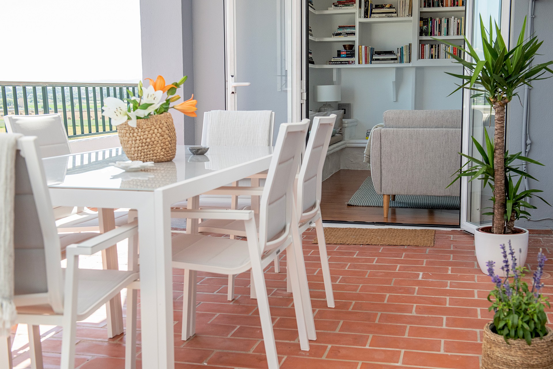 A glass-topped dining table with white chairs on a terracotta-tiled balcony, overlooking mountain views from the apartment.