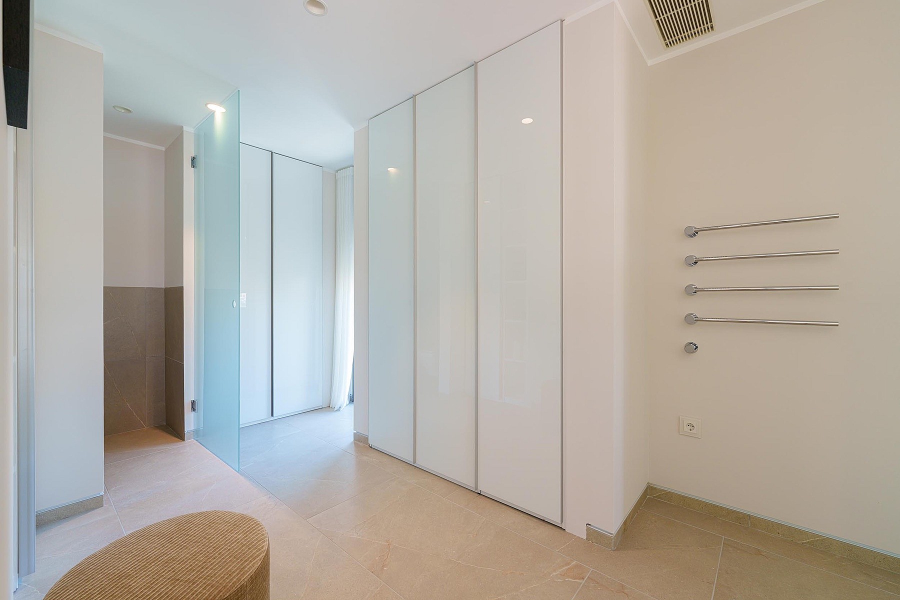 Vestidor luminoso con armarios blancos de suelo a techo, partición de cristal, taburete y toalleros.