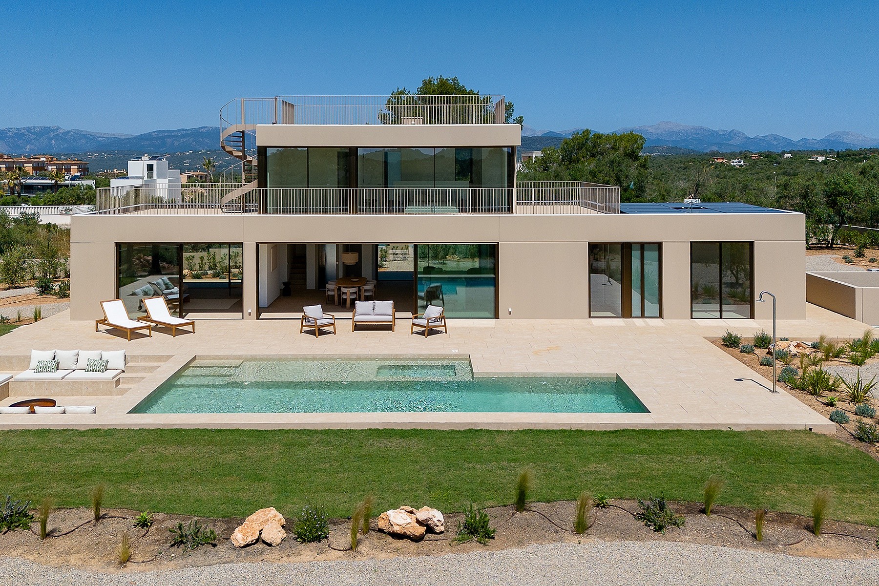 Exterior view of a modern two-story villa with a rectangular swimming pool, stone terrace, and distant mountain backdrop.