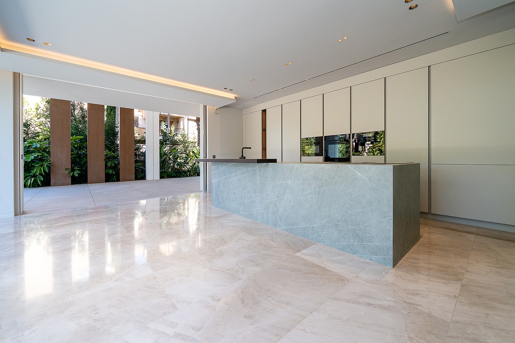 Modern kitchen featuring a large grey stone island, handleless white cabinets, and polished light marble floors. A wide glass door opens to a stone terrace and garden.