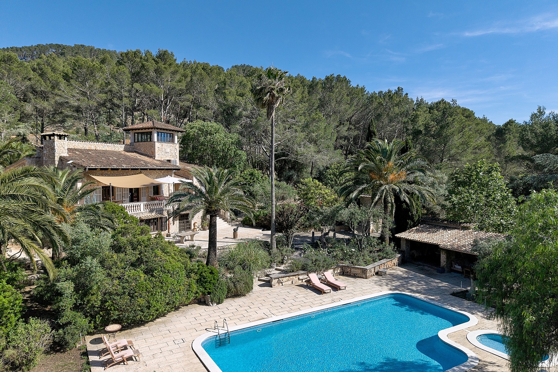 Wide angle view of a stone finca with terracotta roof and a swimming pool in a lush, tree-filled garden in Alaro.