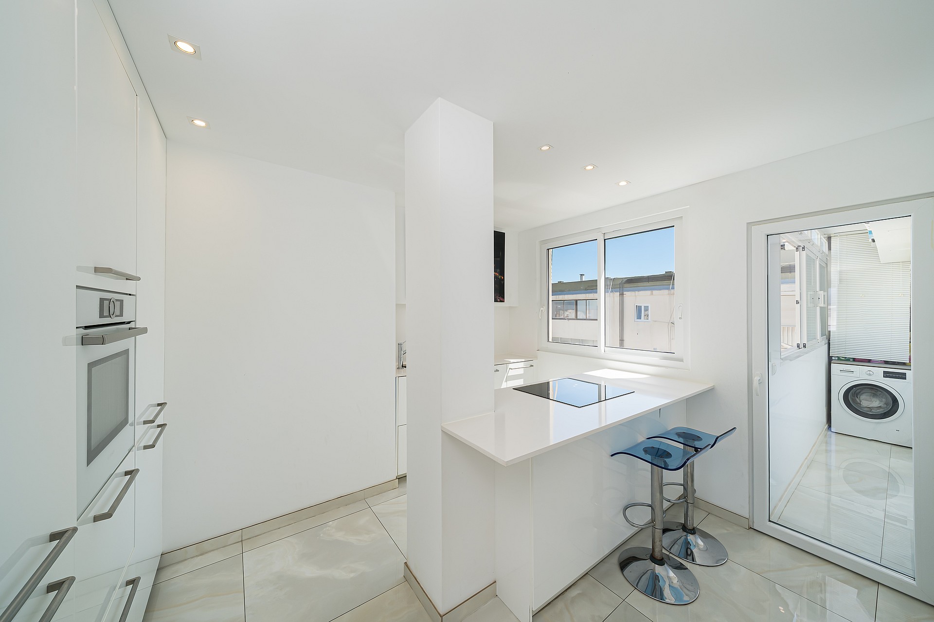 Modern white kitchen featuring a breakfast bar with two blue bar stools, tiled flooring, and an integrated oven.