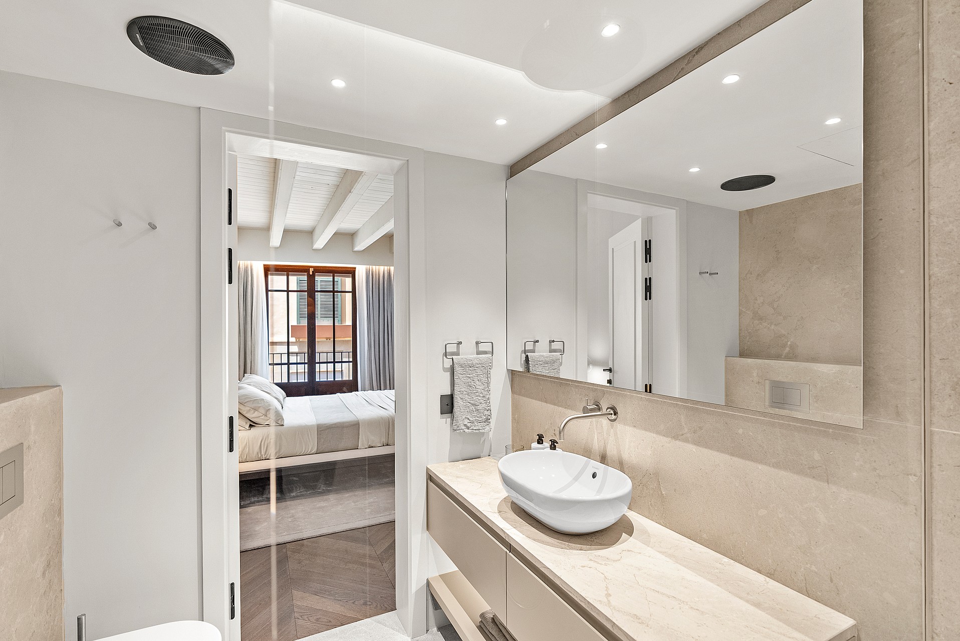 Modern bathroom featuring a stone vanity and white vessel sink, looking into a bedroom with exposed wooden beams.