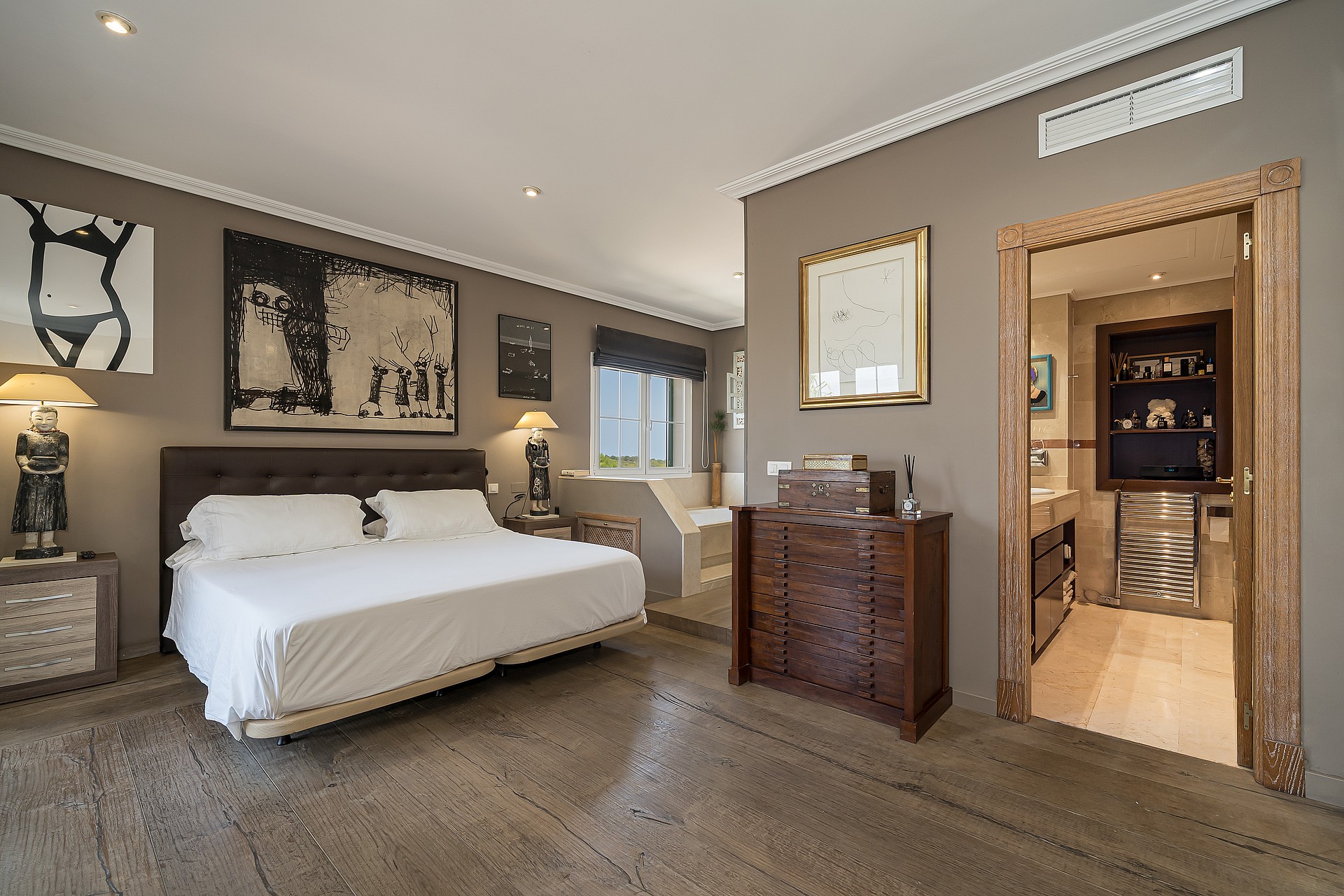 A master bedroom featuring a large bed, dark wooden dresser, and an open doorway leading into a tiled en-suite bathroom.