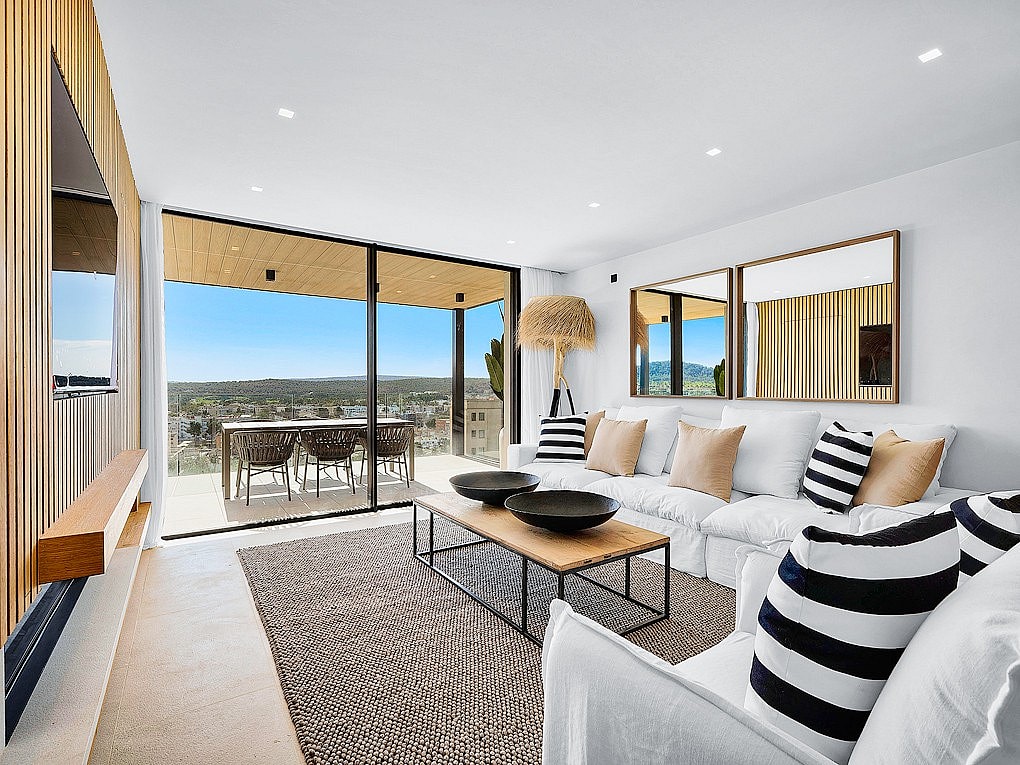 Modern open-plan living room with white sofas, wooden wall paneling, and large glass doors opening onto a terrace with mountain views.