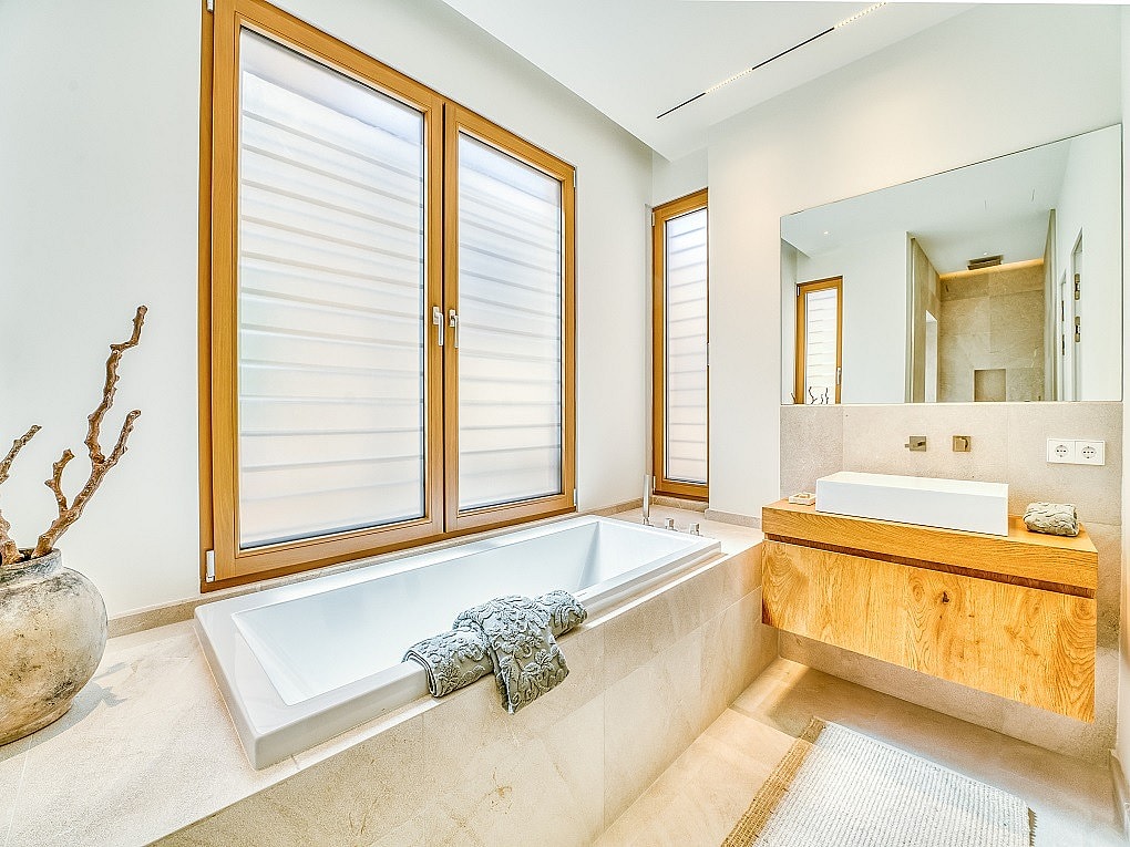 Modern bathroom featuring a rectangular white bathtub, wooden vanity, and large windows with blinds overlooking the city.