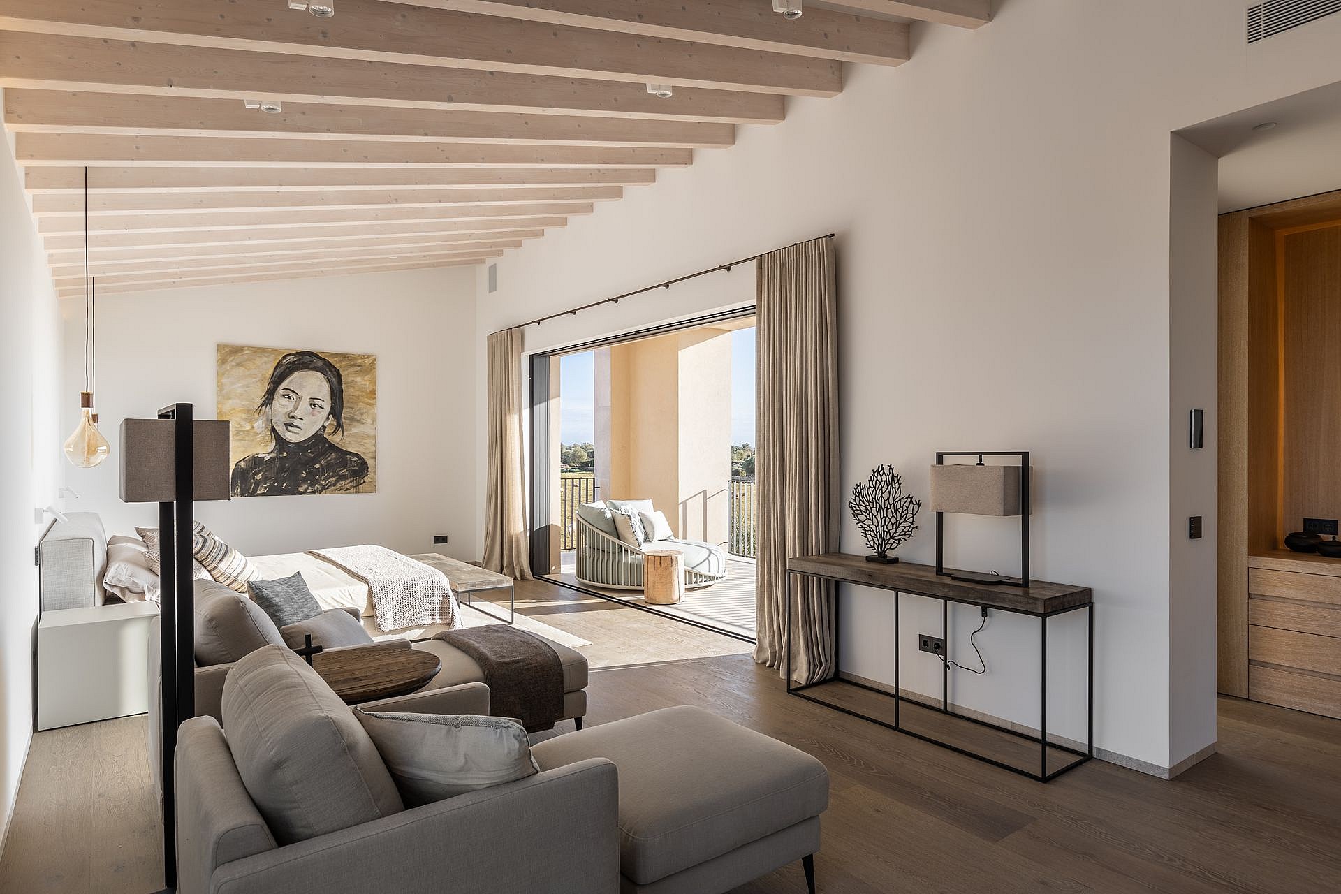 Spacious bedroom with white walls, wooden beam ceiling, gray modular seating, and glass doors opening to a sunlit terrace.