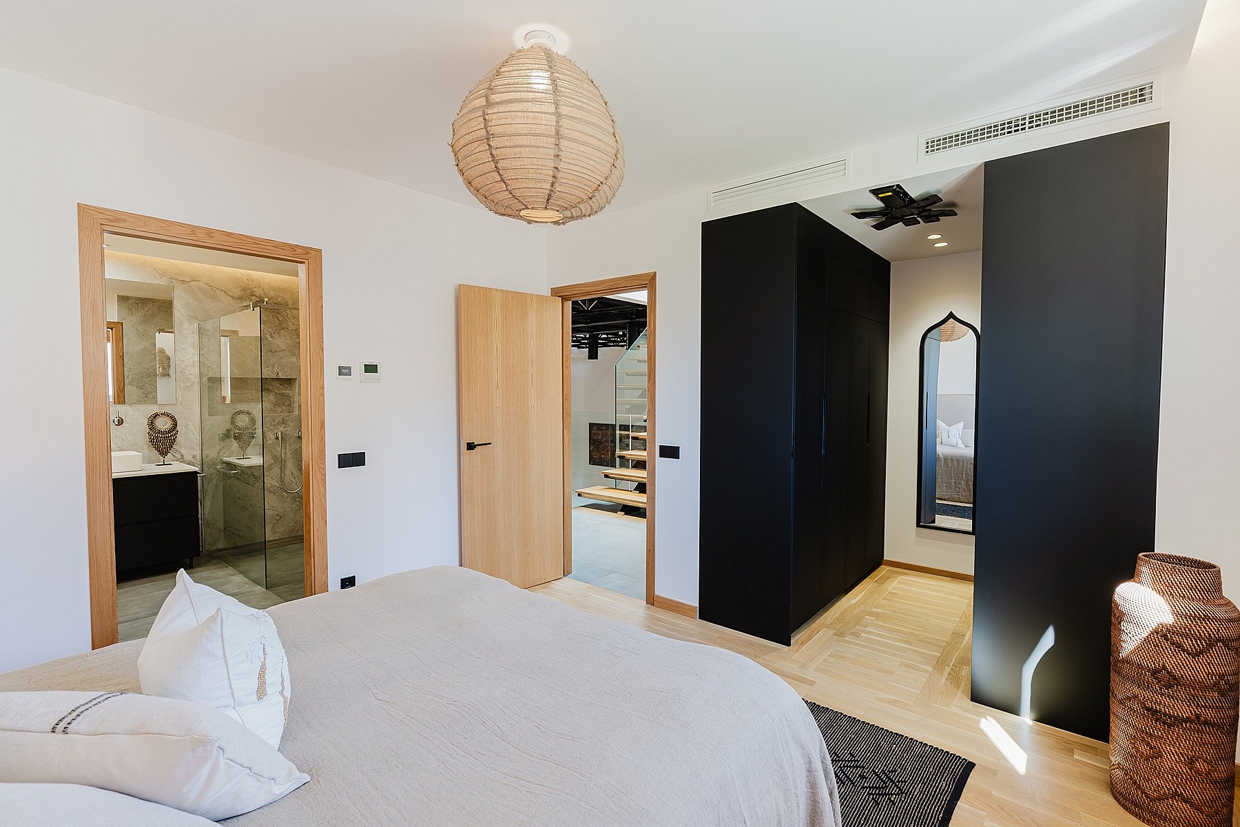 A master bedroom featuring light wood flooring, a beige bedspread, a black wardrobe unit, and an open doorway to a marble bathroom.